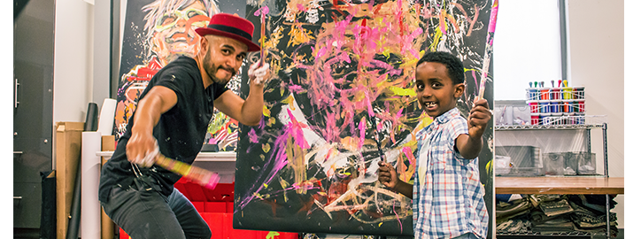 An adult and child artist holding paint brushes in front of a brightly covered canvas